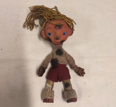 Lalka leży na jasnym tle. Ma żółte, przybrudzone włosy ze sznurków, wełniany swerter, skarpety oraz czerwone szorty i buty. Twarz, dłonie, sweter i lewe kolano są  pobrudzone sadzą. 