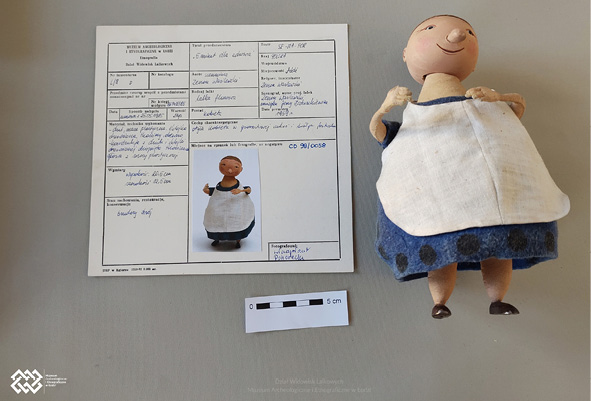 Po lewej stronie leży muzealna karta obiektu z dokładnym opisem lalki gospodyni. Na karcie znajdują się szczegółowe informacje dotyczące wymiarów lalki, autora, stanu zachowania i inne. Lalka jest po prawej stronie karty. Ubrana jest w  niebieską sukienką oraz biały fartuch. Ma namalowany uśmiech, może ruszać głową. Nie widać u niej ruchomych stawów.