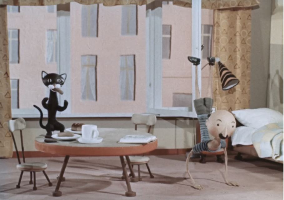 Tłem jest mieszkanie. Po prawej stronie  lalka łysego mężczyzny z czarnym  wąsem stoi na rękach. Ubrany jest w strój do ćwiczeń. Patrzy prosto przed siebie. Po lewej stronie znajduje się mały stolik zastawiony zastawą śniadaniową. Jest na nim otwarta książka. Przy stole  stoją dwa krzesła. Na stoliku stoi na tylnych łpach czarny kot z kiełbasą w pysku i wyciągniętymi w góre przednimi łapkami. 