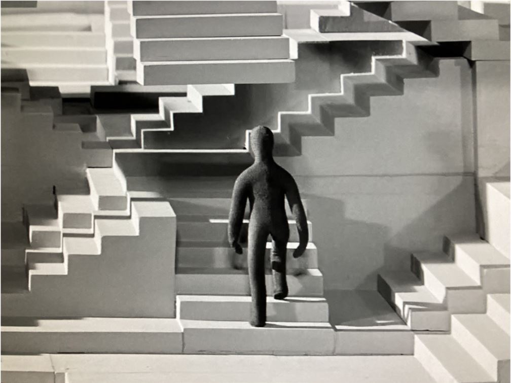 Czarna postać odwrócona tyłem do widza zaczyna wchodzić po schodach. W tle widoczny jest  złożony układ schodów.