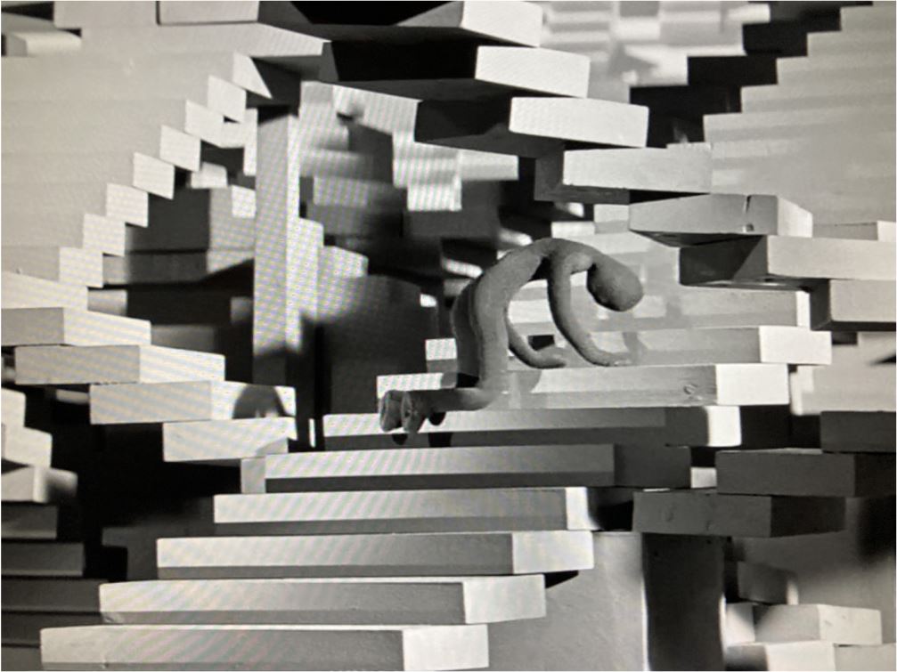 Postać zgięta wpół rękami i nogami opiera się o schody. Wygląda na zmęczoną. W tle złożony układ schodów.