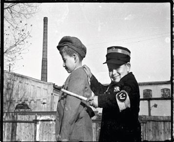 Na czarno-białym zdjęciu widać dwóch chłopców. Chłopiec z lewej strony ma spuszczoną głowę, stoi bokiem do obiektywu, tyłem do chłopca w czarnym mundurze z pałką w ręce. Chłopiec w czarnym mundurze patrzy z uśmiechem w obiektyw. Chłopcy bawią się w policjantów i złodziei. W tle widać komin fabryczny.