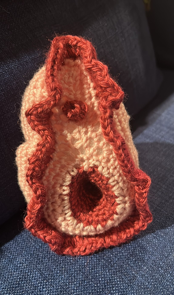 A crocheted vulva by Gill Frigerio, made with orange and brown yarn, lies on a grey sofa. The piece includes a ribbed bright pink section and is crafted with anatomical precision.