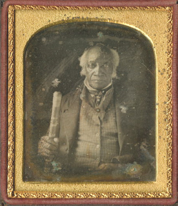 Portret fotograficzny ukazujący elegancko ubranego czarnoskórego mężczyznę w podeszłym wieku. Mężczyzna patrzy prosto w obiektyw.