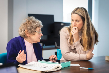 Zdjęcie z przyjaznej wobec wieku biblioteki obrazów przedstawiające dwie kobiety siedzące przy stole konferencyjnym. Starsza tłumaczy coś młodszej, która uważnie słucha.