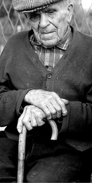 Zdjęcie ukazuje mężczyznę w podeszłym wieku, jego dłonie oparte są na drewnianej lasce. Mężczyzna ma na głowie czapkę, jego wzrok skierowany jest w dół kadru.