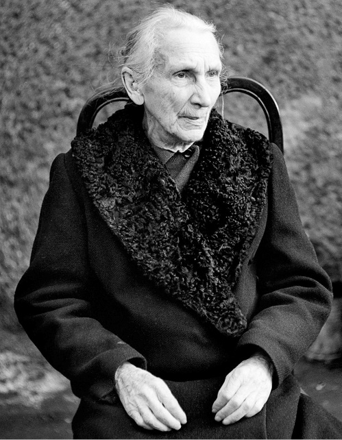 Czarno-biały portret starszej kobiety siedzącej na krześle, ubranej w płaszcz z futrzanym kołnierzem. Kobieta nie patrzy w obiektyw. Zdjęcie emanuje spokojem.