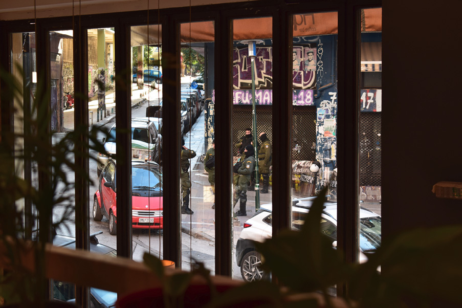 Widok z wnętrza kawiarni przy placu Exarchia w Atenach; przez szybę widać uzbrojonych policjantów w kominiarkach, stojących przy budynku pokrytym graffiti obok zaparkowanych samochodów. 
