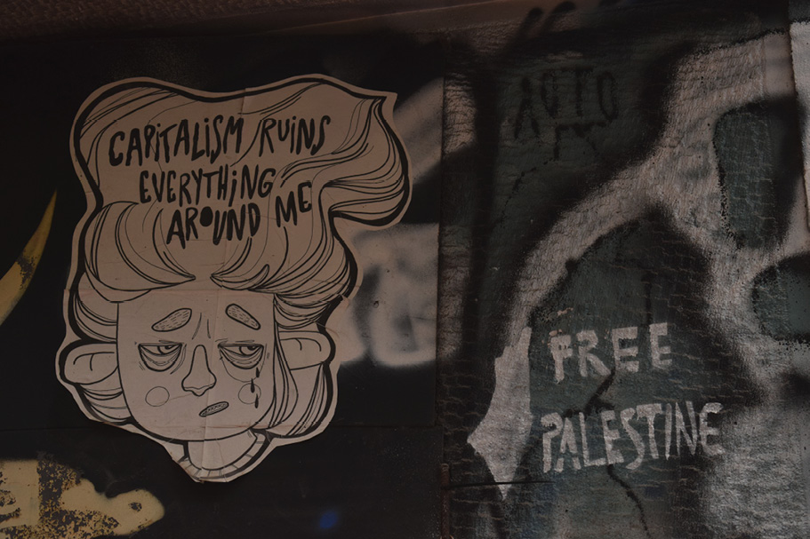Mur z grafiką przedstawiającą zrezygnowaną postać z napisem ‘Capitalism ruins everything around me’; obok hasło ‘Free Palestine’ namalowane sprejem.