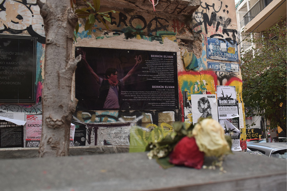 Czarna tablica pamiątkowa poświęcona tureckiemu nastolatkowi zabitemu przez policję; na pierwszym planie czerwona i biała róża, na prawo od tablicy plakaty i graffiti.