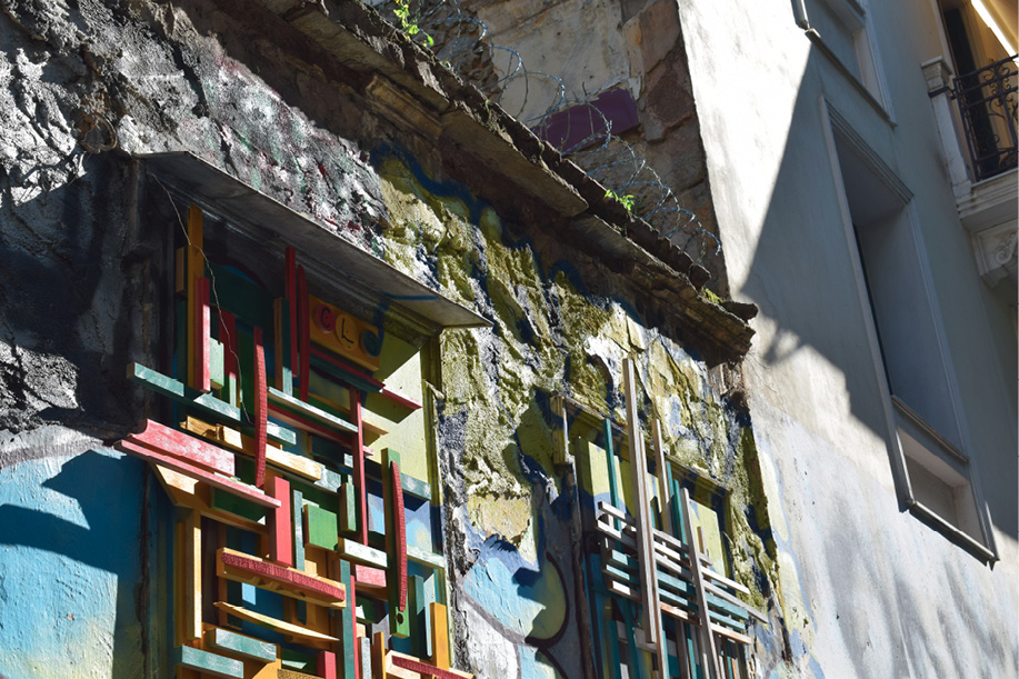 Fragment fasady zniszczonego budynku ozdobionej kolorowymi geometrycznymi instalacjami z małych desek; mur porośnięty mchem, nad nim drut kolczasty.