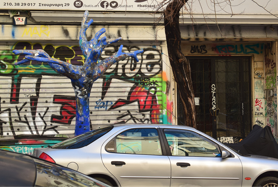 Srebrny samochód zaparkowany przy ulicy; obok niebieska rzeźba drzewa na tle metalowej bramy pokrytej graffiti i kolorowymi napisami.