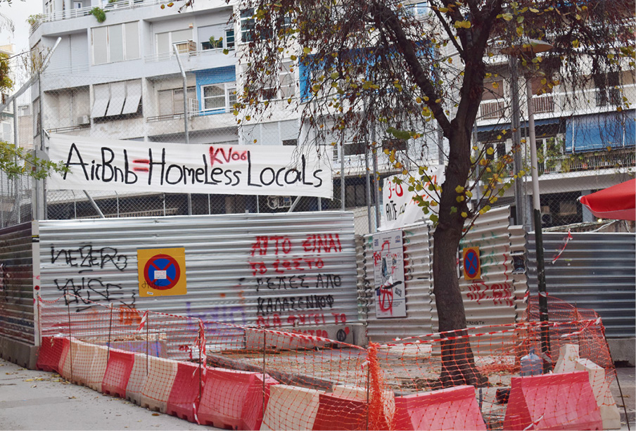 Baner z napisem ‘Airbnb = Homeless Locals’ zawieszony nad ogrodzonym placem budowy w Atenach; metalowe ogrodzenie i mury pokryte są antyturystycznymi hasłami i graffiti.