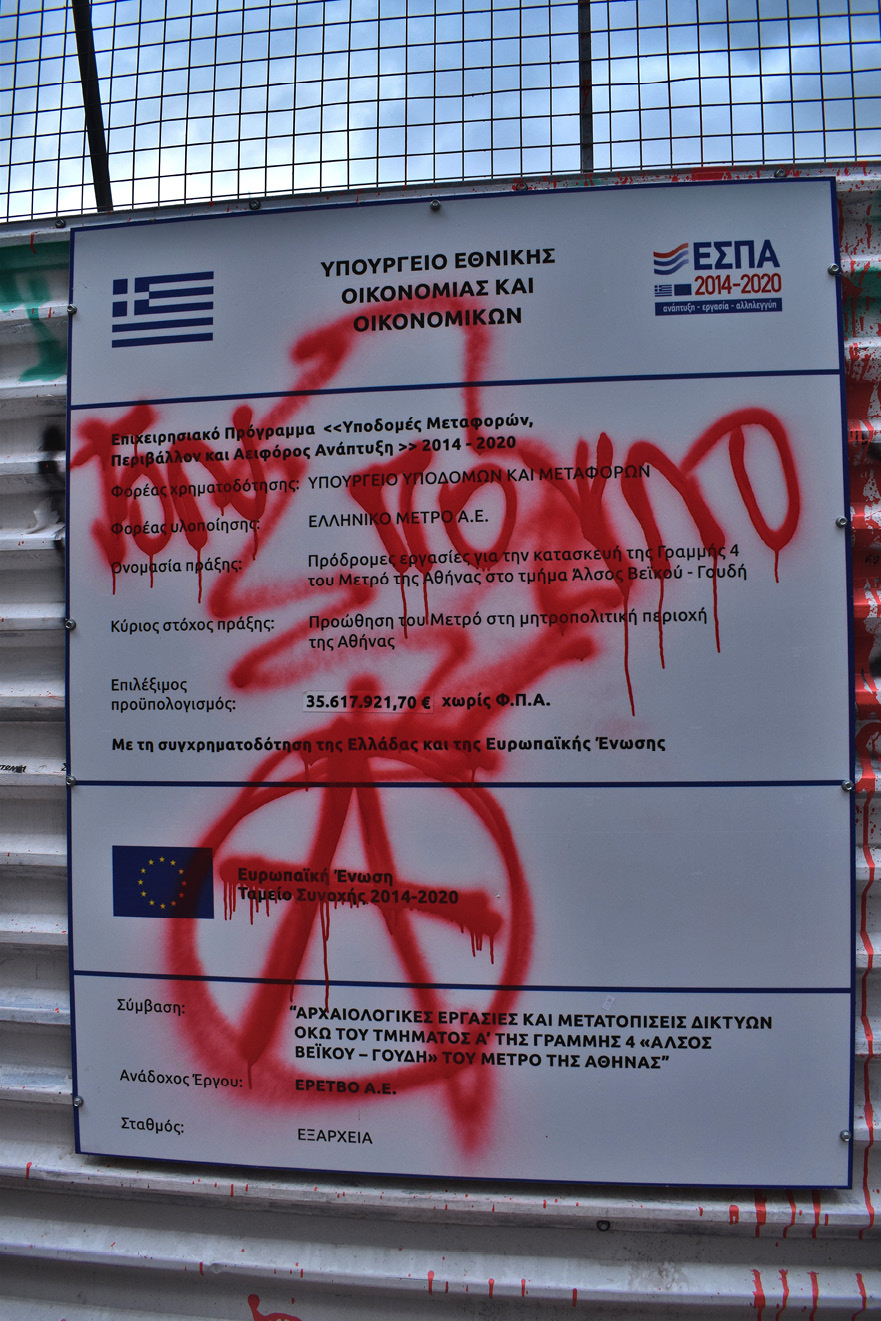 Tablica informacyjna o budowie metra w dzielnicy Exarchia w Atenach, zamazana czerwonym sprejem; widoczny duży znak anarchistyczny.