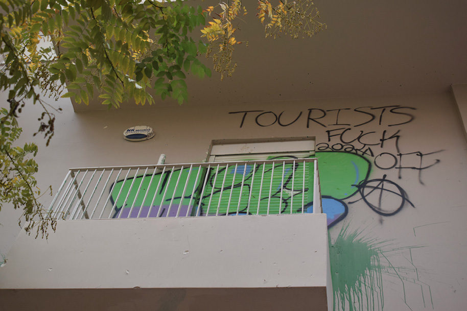 Widok na balkon w Atenach z graffiti i napisem ‘Tourists fuck off’; obok hasła widoczny znak anarchistyczny i kolorowe malunki na ścianie oraz rozpryskana zielona farba.