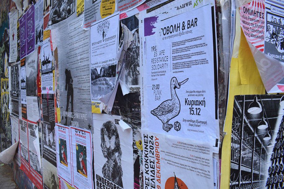 Zbliżenie na ścianę w dzielnicy Exarchia w Atenach, pokrytą warstwami plakatów z hasłami politycznymi, ogłoszeniami o wydarzeniach i grafikami o anarchistycznym charakterze.