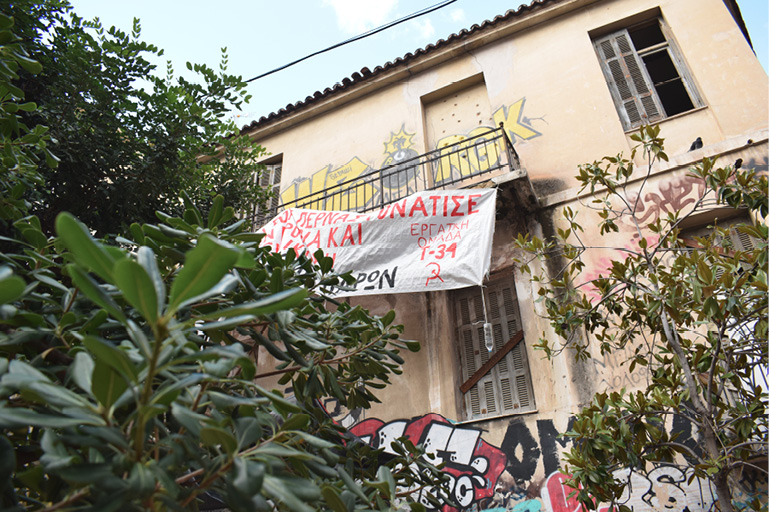 Opuszczony budynek w Exarchii z balkonem, na którym zawieszono transparent z hasłem po grecku; zniszczona elewacja pokryta graffiti, otoczona gęstą roślinnością. 