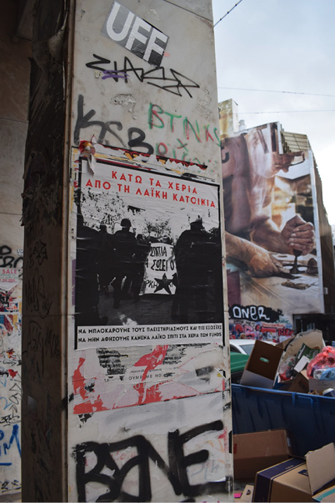 Filar budynku w Exarchii pokryty graffiti i plakatem przedstawiającym interwencję policji podczas zamieszek. W tle mural przedstawiający człowieka pracującego z drewnem, obok kontenery na odpady.