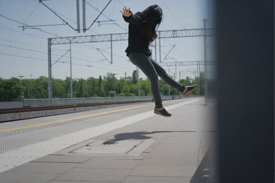 A person with long hair, mid-air in a jumping pose, faces away from the camera on an empty railway platform. Part of the image is obscured by a dark vertical object on the right. Tracks, overhead wires, and trees are visible in daylight.