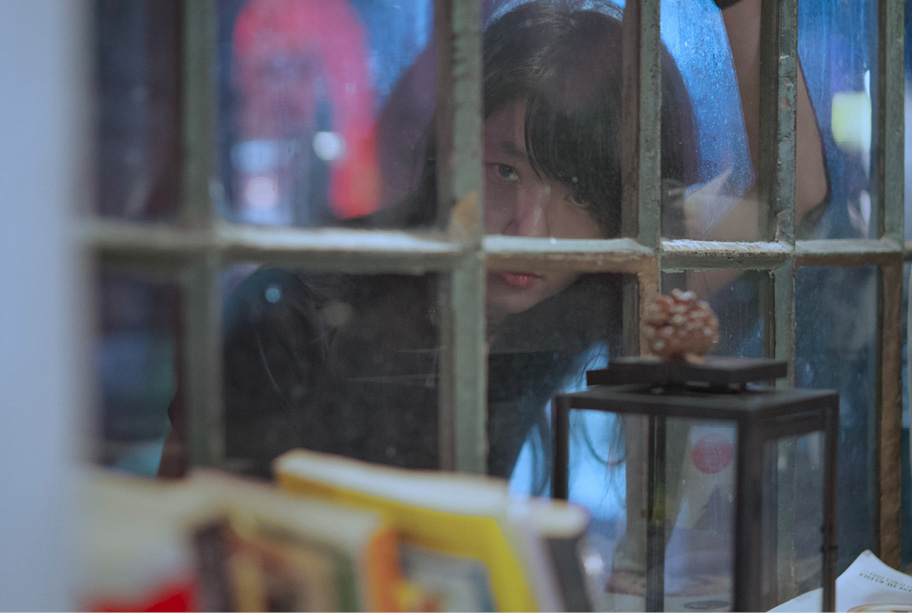 Seen through a multi-paned window, a person leans forward with part of their face obscured by the frame. A lantern, pine cone, and blurred books are visible indoors in the foreground. Streetlights are faintly visible in the background.