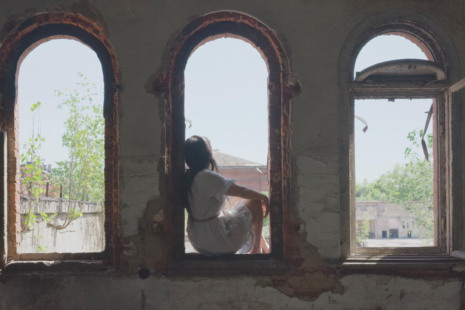 A person in a white dress, back to the camera, sits on a windowsill in a dark, dilapidated industrial-style room with three arched windows. The walls are cracked and peeling. Outside, trees and buildings are visible under a clear sky.