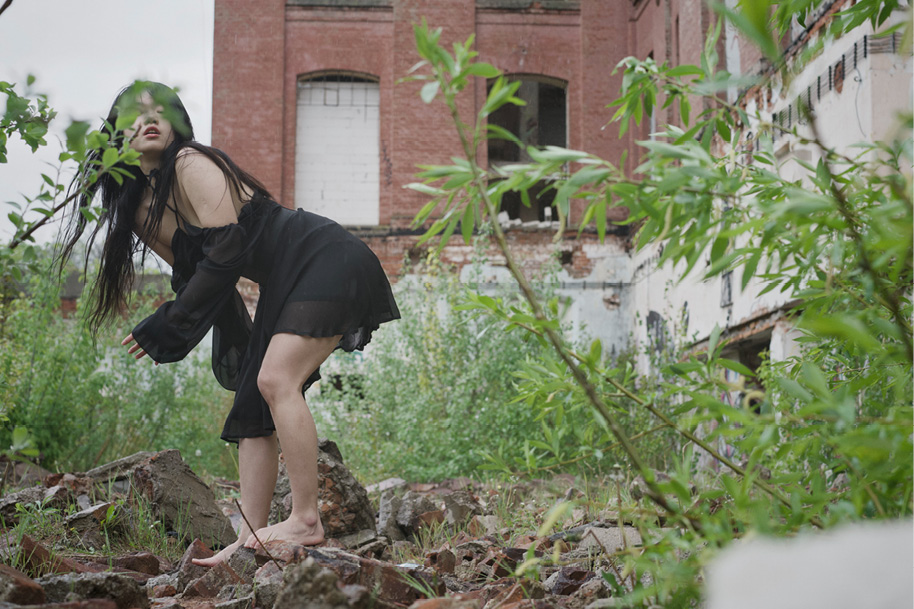 A person in a black dress crouches on rocky ground, surrounded by green plants and trees. Behind them stands a dilapidated brick building with broken windows. Her face is partially obscured by some leaves.