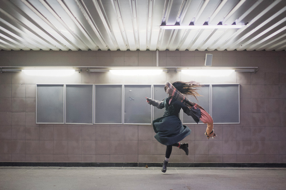 A person in a dark coat and pink scarf jumps mid-air in an underground passage with a tiled ceiling and walls. Lights are mounted above, and empty poster boards are on the wall. Her face is obscured by the moving scarf.