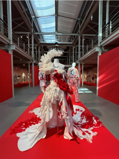 The "Arkadius. Powerful Emotions. Confrontations" exhibition hall with industrial steel structures and a red floor, showcasing a dramatic, asymmetrical long train white gown by Arkadius, with vibrant white and red feather details. 