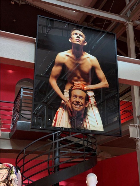A large-scale, the iconic fine art photograph of designer Arkadius by Robert Wolański, displayed in a exhibition space above a spiral staircase. The photograph shows a shirtless designer looking upward while holding a smiling, bloody replica of his head.