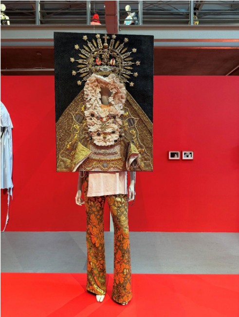 An avant-garde look from the "Virgin Mary Wears the Trousers" collection by Arkadius at his retrospective museum exhibition. A mannequin features patterned gold and an oversized rectangular canvas as the torso, depicting a detailed religious icon of a crowned Madonna with intricate ruffles and gold embroidery.