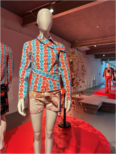 A white faceless mannequin wearing a lace cap displays an avant-garde outfit from the Paulina” collection by Arkadius at his fashion exhibition. The styling includes a fitted, asymmetric long-sleeve shirt with a dense red and blue motives inspired by Kurpie paper-cutouts, paired with light beige shorts featuring heavily frayed red edges. The background shows the exhibition space with a red platform, a decorative branch arrangement, and other mannequins.