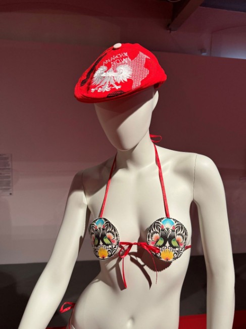 A female mannequin displayed at the exhibition, wearing a red flat cap with a white eagle and the text "ANARCHY NOW", paired with a bikini top featuring colourful Łowicz folk roosters from “Paulina” collection by Arkadius. 