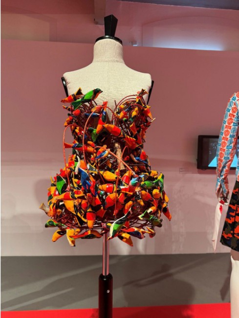 An avant-garde strapless bird dress designed by Arkadius for “Paulina” collection, displayed on a mannequin at his retrospective exhibition. The dress is crafted from densely arranged, brightly painted wooden bird figures in shades of red, yellow, and green.