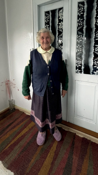 Pani Zenaida Aleksiejuk stoi w domu na ręcznie tkanym chodniku. Fotografia przedstawia rozmówczynię oraz przykład tekstyliów użytkowanych w przestrzeni domowej.