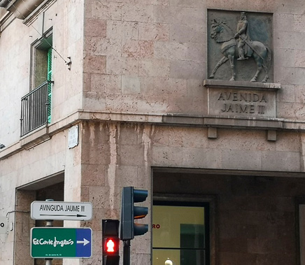 A street corner, with a lamppost-mounted sign in the bottom-left of the image, and a stone sign in relief on a wall in the top-right corner. 