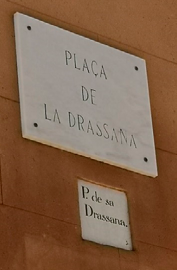 A marble street sign mounted on a wall, above a smaller ceramic sign set into the wall. 