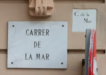 A marble street sign mounted on a wall, to the left of a smaller ceramic sign set into the wall. 