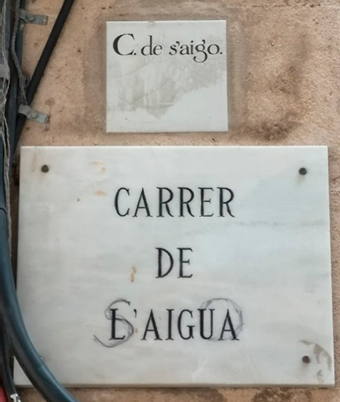 A marble street sign mounted on a wall, below a smaller ceramic sign set into the wall. 