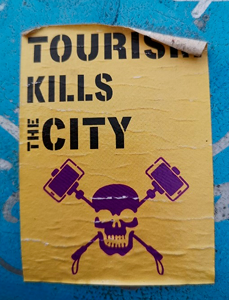 An anti-tourist sticker stuck on a blue pillar box, with the top right corner peeling down. 