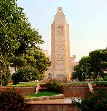 A tall stone-built monument, featuring writing in relief, surrounded by plants and water features.  