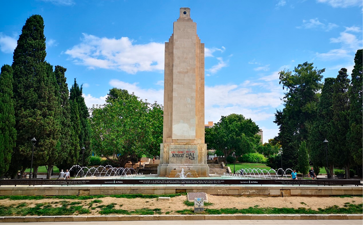 A tall stone-built monument, but with the writing in relief removed. It is surrounded by plants, water features, and an iron plaque featuring an inscription. 
