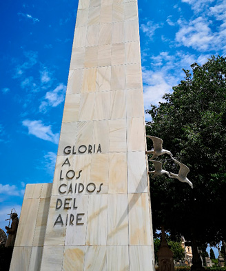 A tall and narrow marble-clad monument, featuring an inscription in metal lettering, and sculptures of birds plummeting on the right. 
