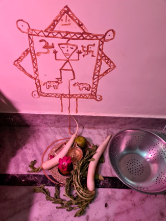 On the left, Lord Vishnu represented on a wall through red ochre with offerings including radishes, flowers, and fruits placed on a plate below it. 