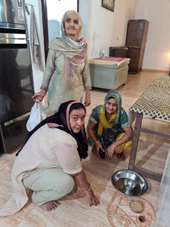 On the left, three women in traditional Haryanvi attire participating as two women are sitting near a floor decoration made of red ochre, with a steel plate and bowl beside them, while an older woman stands behind them. 
