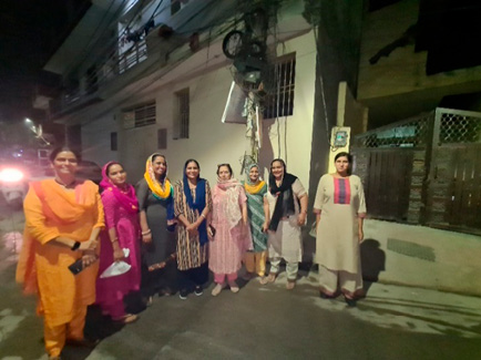 On the right, a group of eight Haryanvi women standing together on a residential street at night in Gurugram, dressed in colourful traditional Haryanvi attire, smiling and posing for the photo.