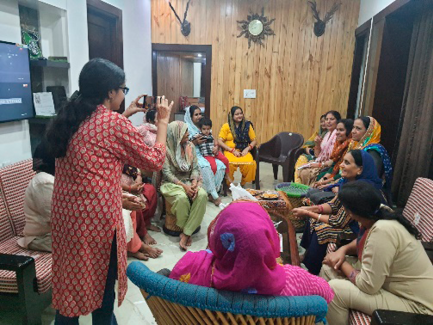 On the left, a group of Haryanvi women gathered indoors for the Dev Uthan Ekadashi celebration, some seated and chatting while one woman (the author) photographs the gathering. 