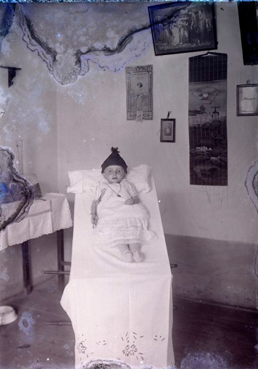 Dead baby with open eyes, wearing a white dress, knitted shoes, a chain with a medal, and a triangular bonnet. Its left hand is on its belly, and it holds a lily in its right hand. Its head rests on a pillow, and its body lies on an inclined litter, covered with impeccably embroidered white linen cloth.