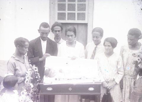 A small baby with clasped hands lies on a coffin placed on a table outside a house. Seven adults (three men and four women) and two girls surround the coffin. Two women, one on each side, hold modest wreaths. Five adults look at the baby with grief, while a couple on the right have smiling expressions.